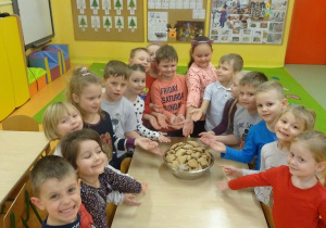 Grupa uśmiechniętych dzieci stoi wokół stołu, rączkami wskazują miskę wypełnioną upieczonymi pierniczkami.